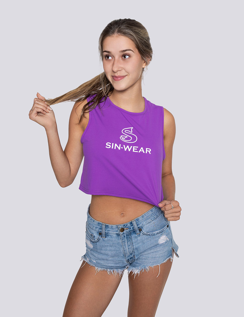 Woman wearing a purple 'Sin-Wear' tank top and denim shorts on a plain background